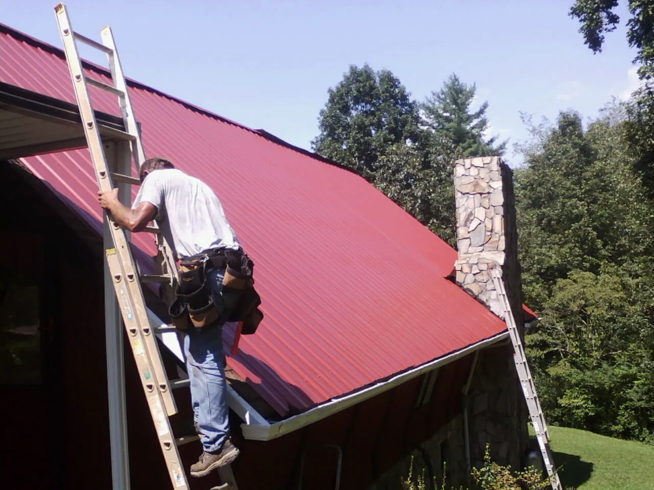 Roofer on a ladder beside a red metal roof and stone chimney — Soffit & Fascia Repair by Roofing Brothers in Hudsonville